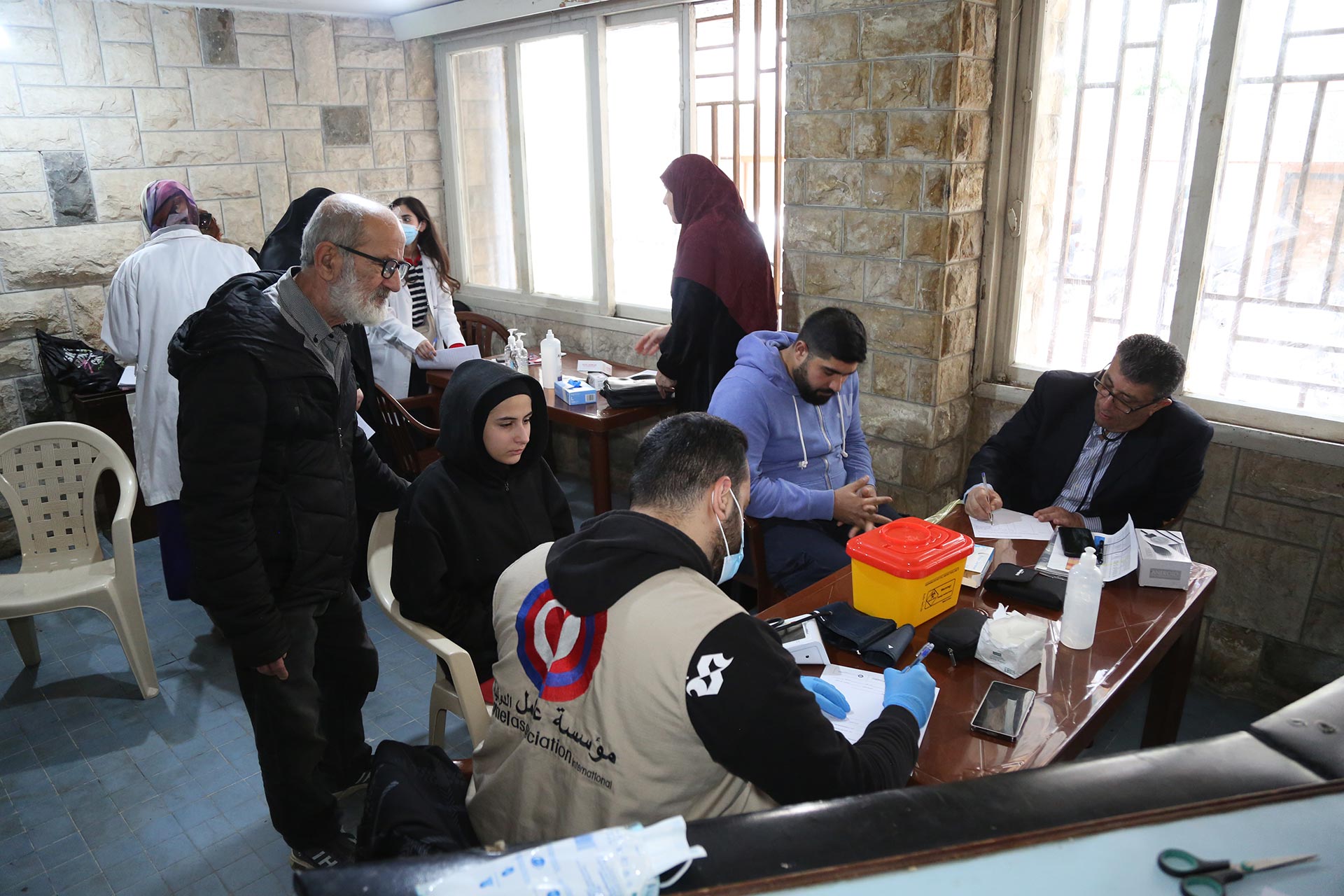 Health workers assist people at several desks inside a clinic room, with medical supplies and paperwork on the tables
