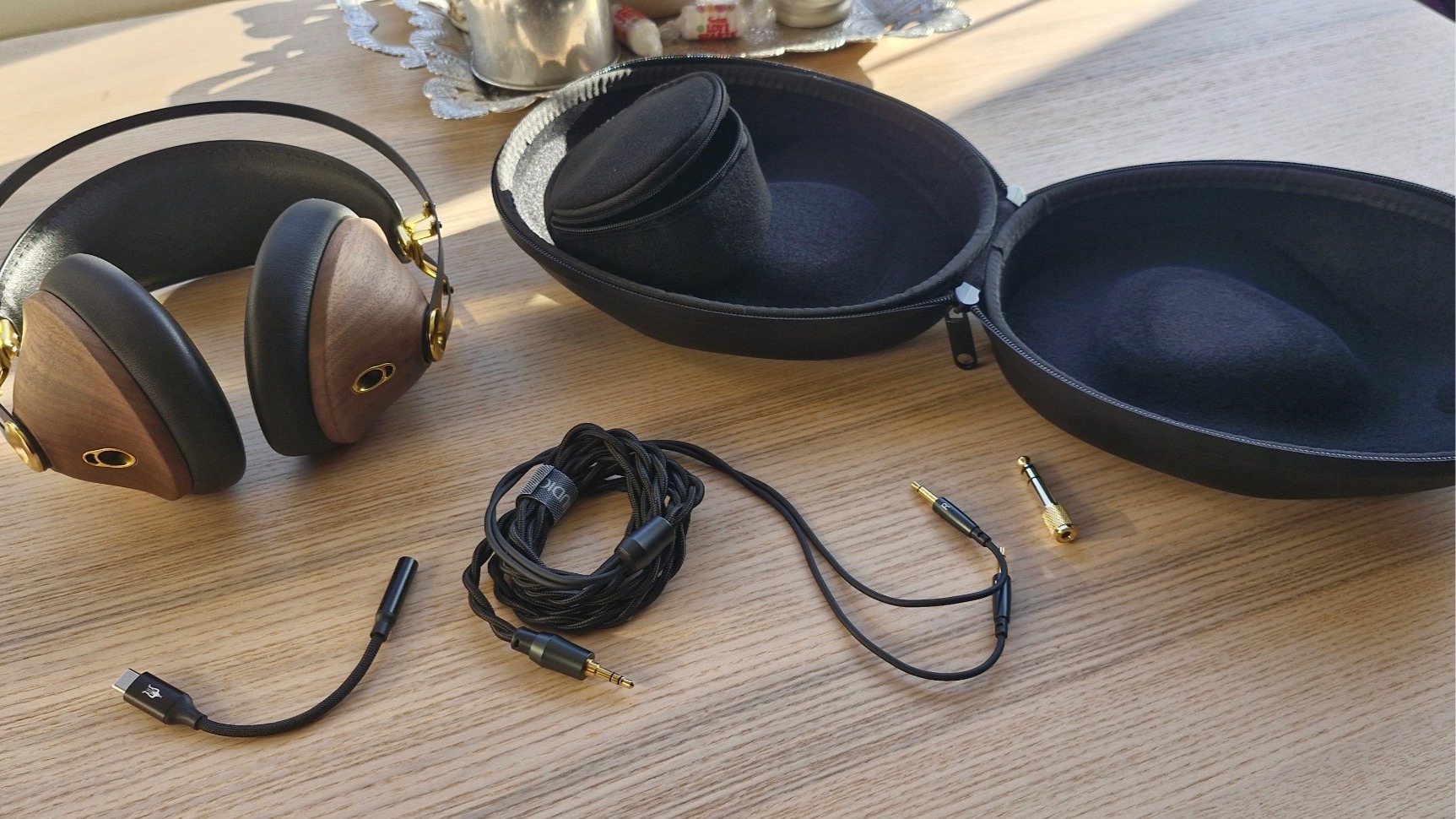 Meze Audio 99 Classics 2nd Gen over-ear headphones placed on a wooden table, alongside their hard carry case, provided cables, adapters and mini DAC.