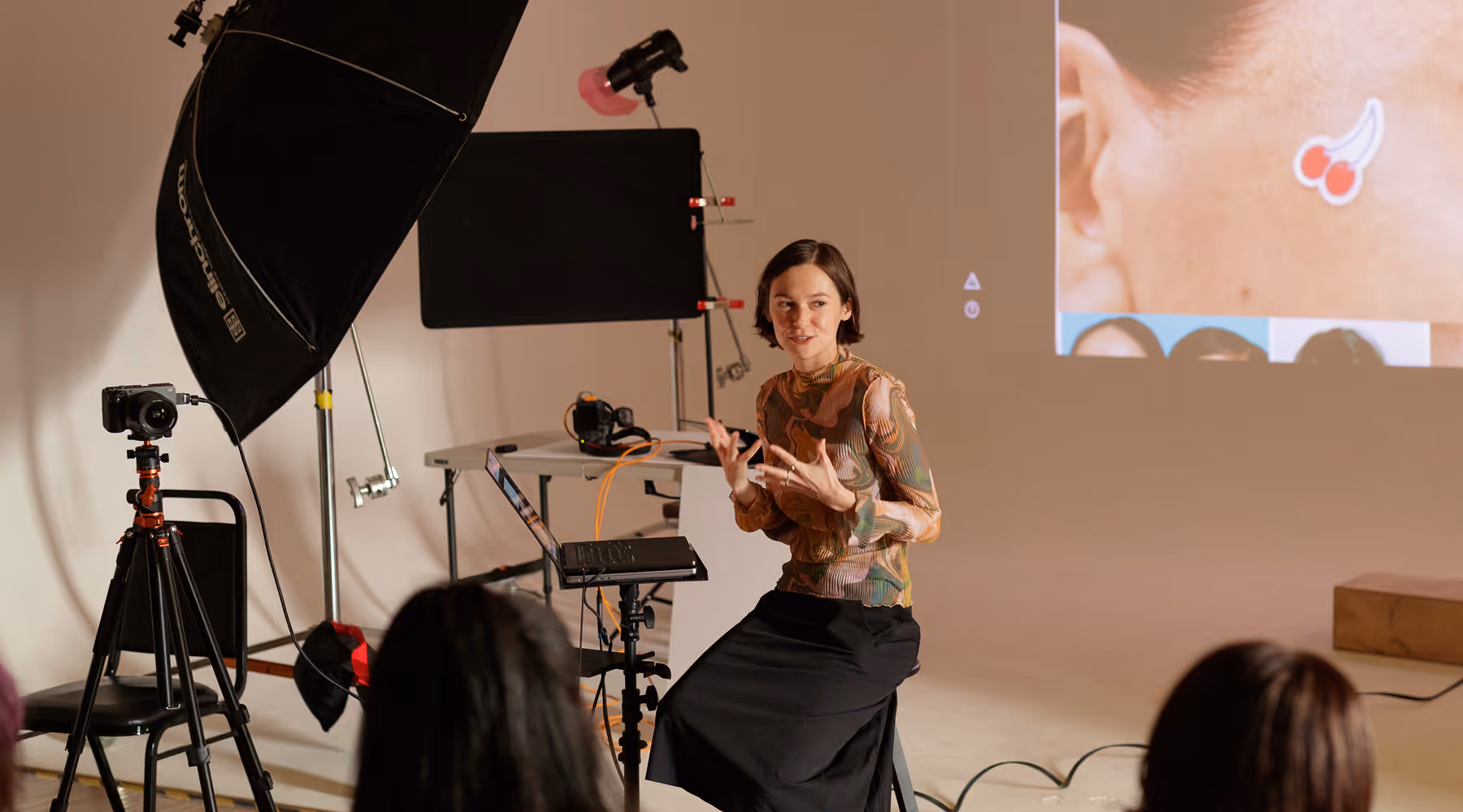 A presenter sitting on a stool gesturing with hands while speaking in front of a photography workshop