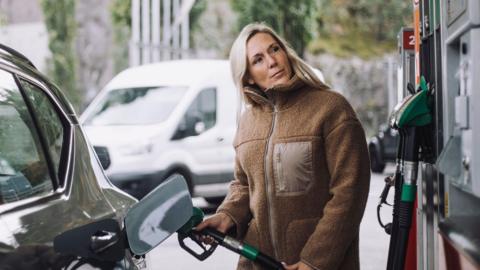 A stock photo of lady in a brown jacket filling up her car with fuel at a petrol station.