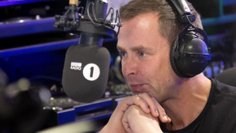 Scott Mills leaning into the microphone in the Radio 1 studio with headphones on.